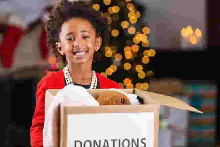 A smiling child in a red sweater holds a box labeled DONATIONS filled with toys, including a teddy bear, as festive holiday lights twinkle in the background—supporting the Nanaimo Brain Injury Society’s mission to spread joy.