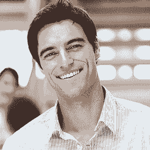 A man with short dark hair and a striped shirt smiles at the camera in a brightly lit indoor setting, representing the Nanaimo Brain Injury Society, with blurred lights and a person in the background.