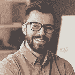 A man with dark hair, a beard, and glasses smiles while sitting indoors. Wearing a button-up shirt in an office setting, he appears engaged in Brain Injury Support work at NBIS.