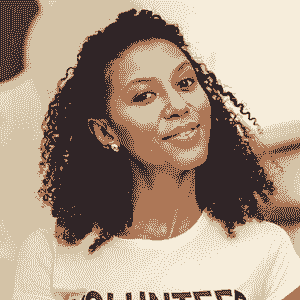 A woman with curly hair smiles at the camera, wearing a shirt that partially shows the word VOLUNTEER, representing NBIS and Brain Injury Support. The background is blurred.