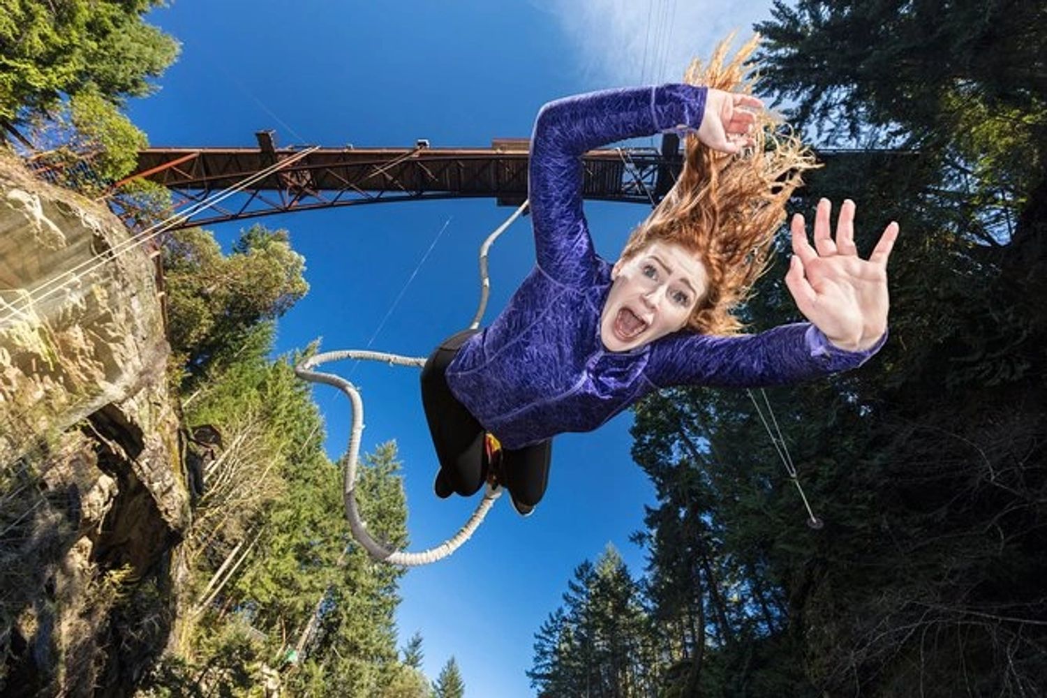 A person with long hair and a purple top is bungee jumping from a bridge, looking surprised and reaching out, with trees and a clear blue sky in the background—highlighting the thrill and risks that organizations like Nanaimo Brain Injury Society address.