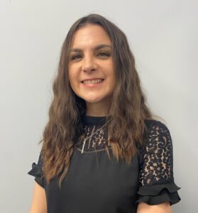 Angie Bisset, with long wavy brown hair and a black lace short-sleeved top, stands smiling in front of a light gray wall.