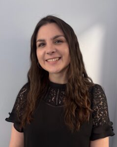 Angie Bisset, a woman with long brown hair, smiles while wearing a black lace short-sleeve top and standing against a light gray background.