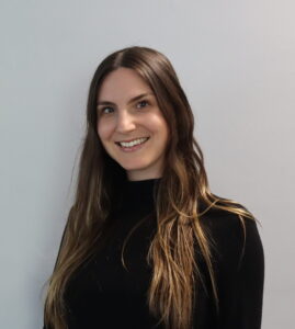 A woman with long brown hair, wearing a black top, stands smiling in front of a plain light gray background, embodying the strength and hope inspired by Brain Injury Support.