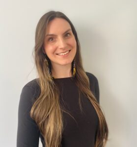 Camilla Arnold, a woman with long brown hair wearing a black top and brown pants, stands against a plain light-colored wall, smiling at the camera.
