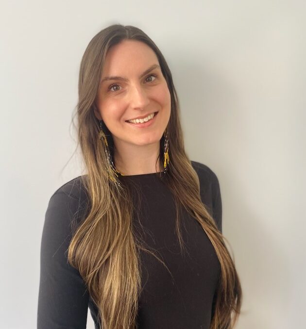 Camilla Arnold, a woman with long brown hair wearing a black top and brown pants, stands against a plain light-colored wall, smiling at the camera.