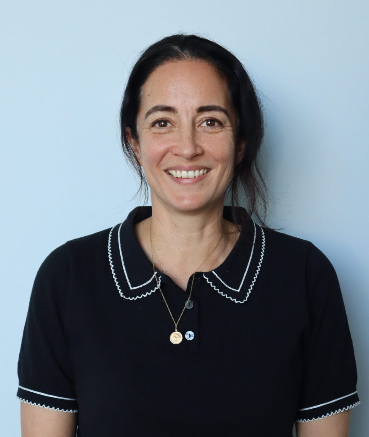 Adrienne Bennest, with long dark hair, wears a black short-sleeve top with white trim, blue jeans, and a white watch as she stands smiling in front of a light blue wall.