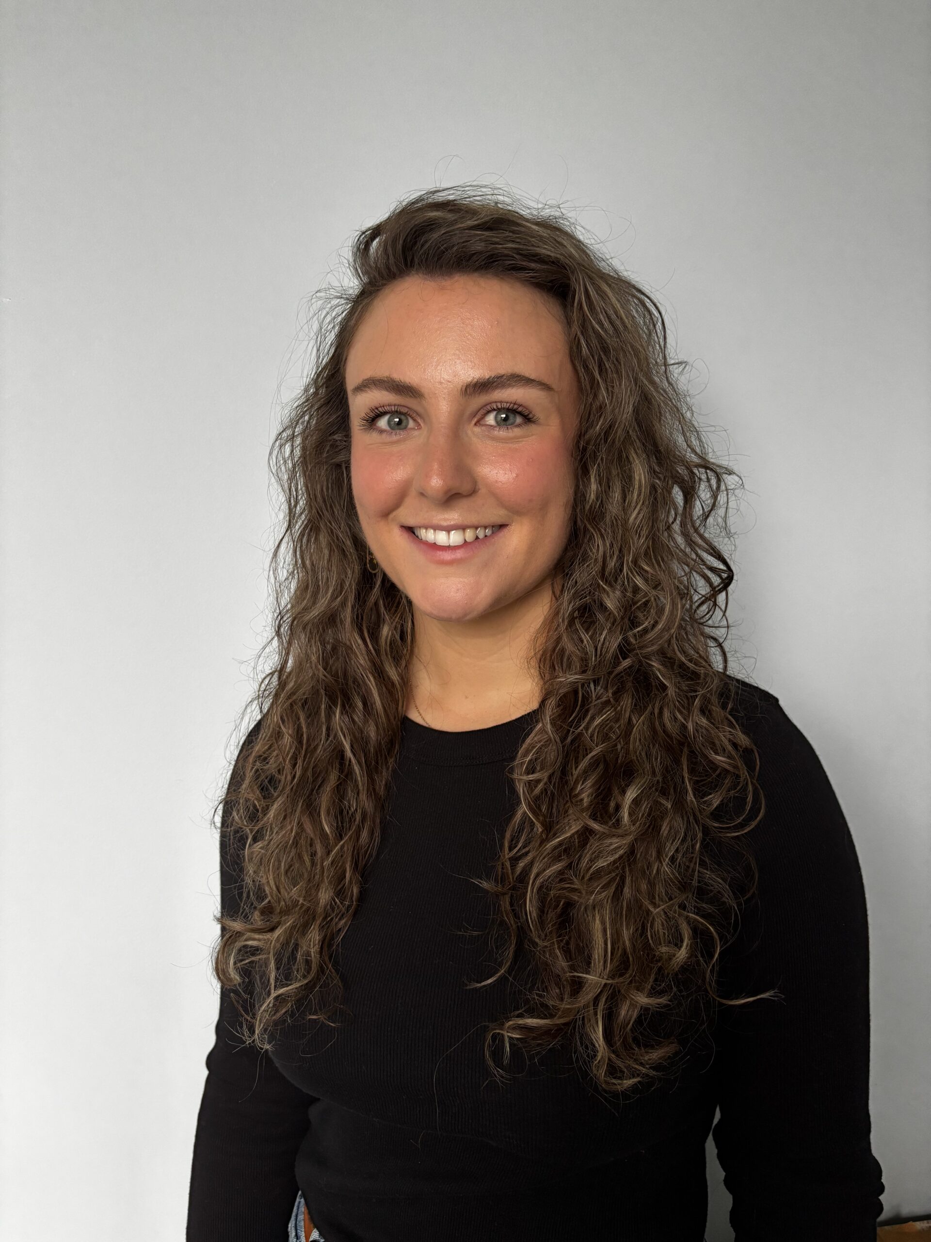 Jess Johnson, a woman with long, curly brown hair and light eyes, smiles at the camera. She is wearing a black long-sleeve top and stands against a plain light gray background.