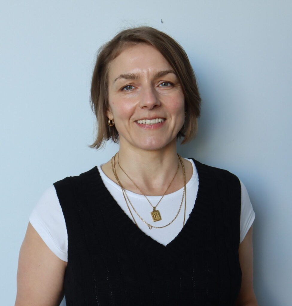 A woman with short brown hair smiles at the camera, wearing a white t-shirt under a black sleeveless sweater and layered gold necklaces, standing against a plain light blue wall.