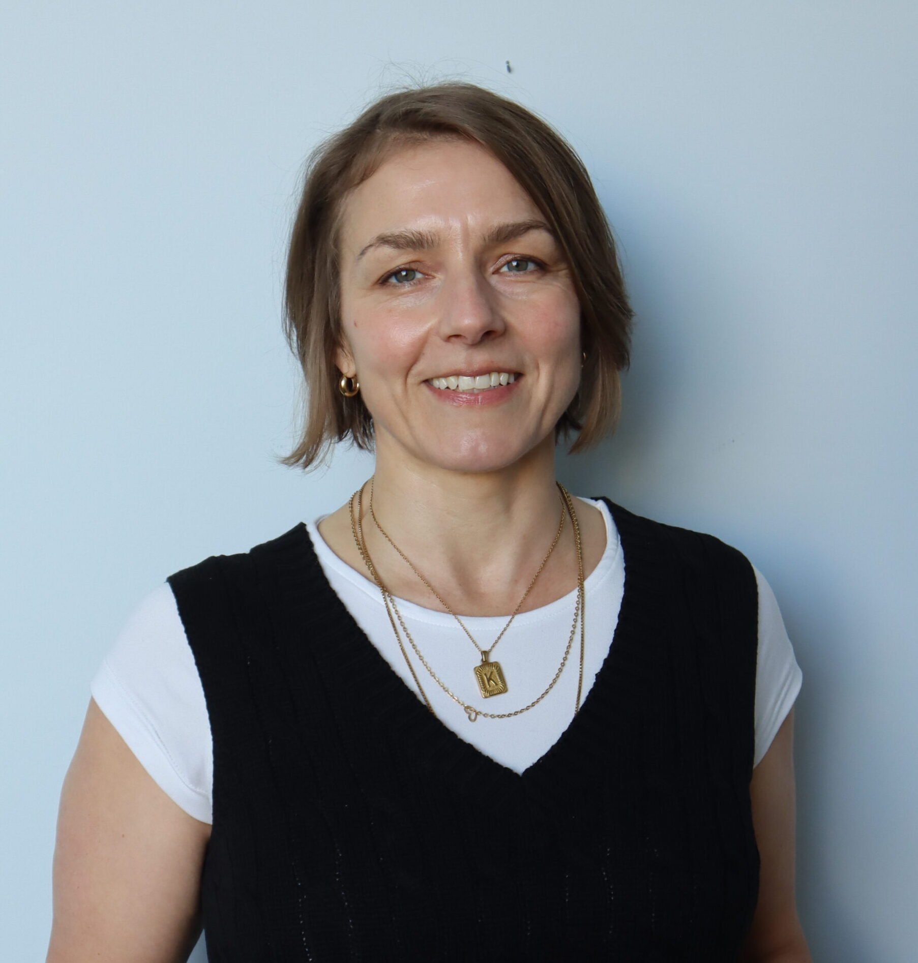 A woman with short brown hair smiles at the camera, wearing a white t-shirt under a black sleeveless sweater and layered gold necklaces, standing against a plain light blue wall.