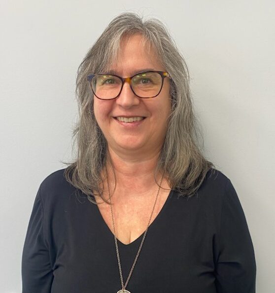 Marianne Verigin, a woman with gray hair and glasses, smiles at the camera in a black top and long necklace, standing against a plain light gray wall.