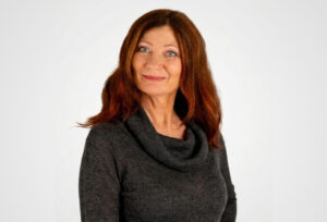 Martina Holblingova, a woman with long reddish-brown hair, stands in front of a plain white background wearing a dark cowl-neck sweater. She smiles gently with one hand on her hip.