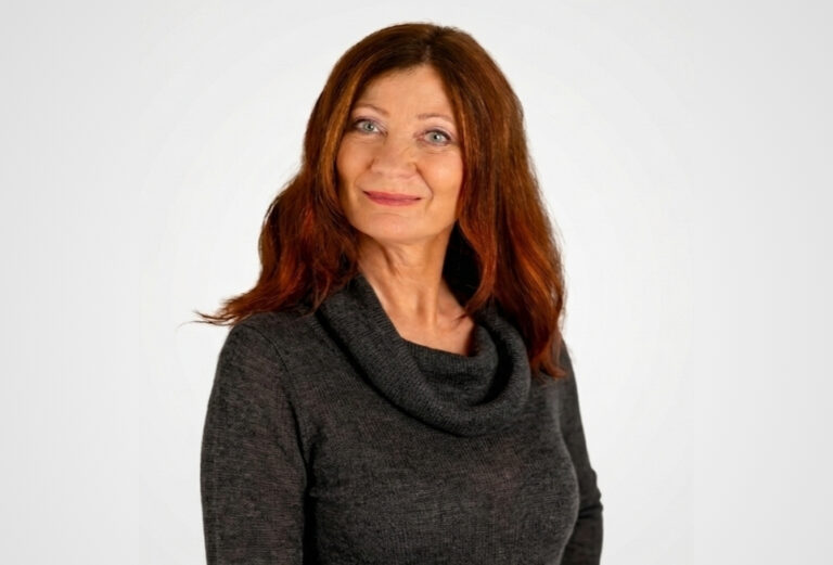 Martina Holblingova, a woman with long reddish-brown hair, stands in front of a plain white background wearing a dark cowl-neck sweater. She smiles gently with one hand on her hip.