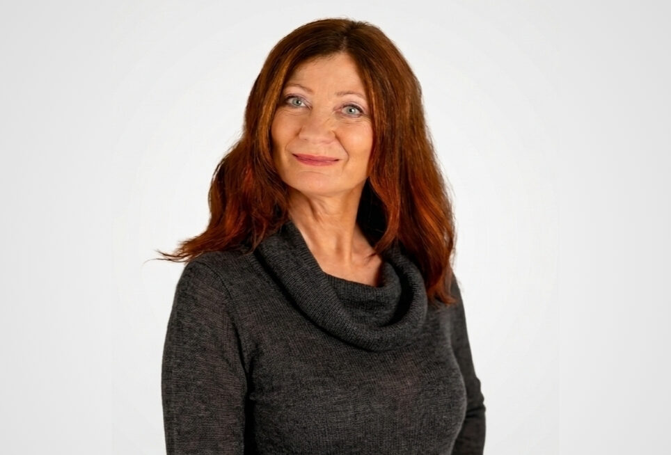 Martina Holblingova, a woman with long reddish-brown hair, stands in front of a plain white background wearing a dark cowl-neck sweater. She smiles gently with one hand on her hip.
