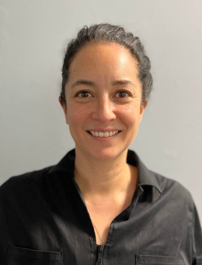 A person with dark hair tied back, wearing a black collared shirt, smiles at the camera in front of a plain light gray background, representing the Nanaimo Brain Injury Society's commitment to Brain Injury Education.