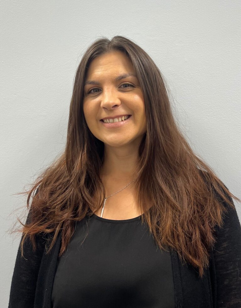 A woman with long brown hair wearing a black top and cardigan stands against a plain light gray wall, smiling at the camera, representing the Nanaimo Brain Injury Society and its commitment to supporting those affected by brain injury.