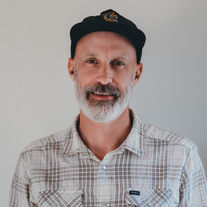 A man with a gray beard and mustache wearing a black cap and a light plaid shirt stands in front of a plain light background, smiling slightly, representing NBIS and Brain Injury Education.