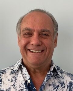A smiling older man with short gray hair wearing a white and blue floral print shirt stands in front of a plain light-colored background, representing NBIS and its commitment to Brain Injury Education.
