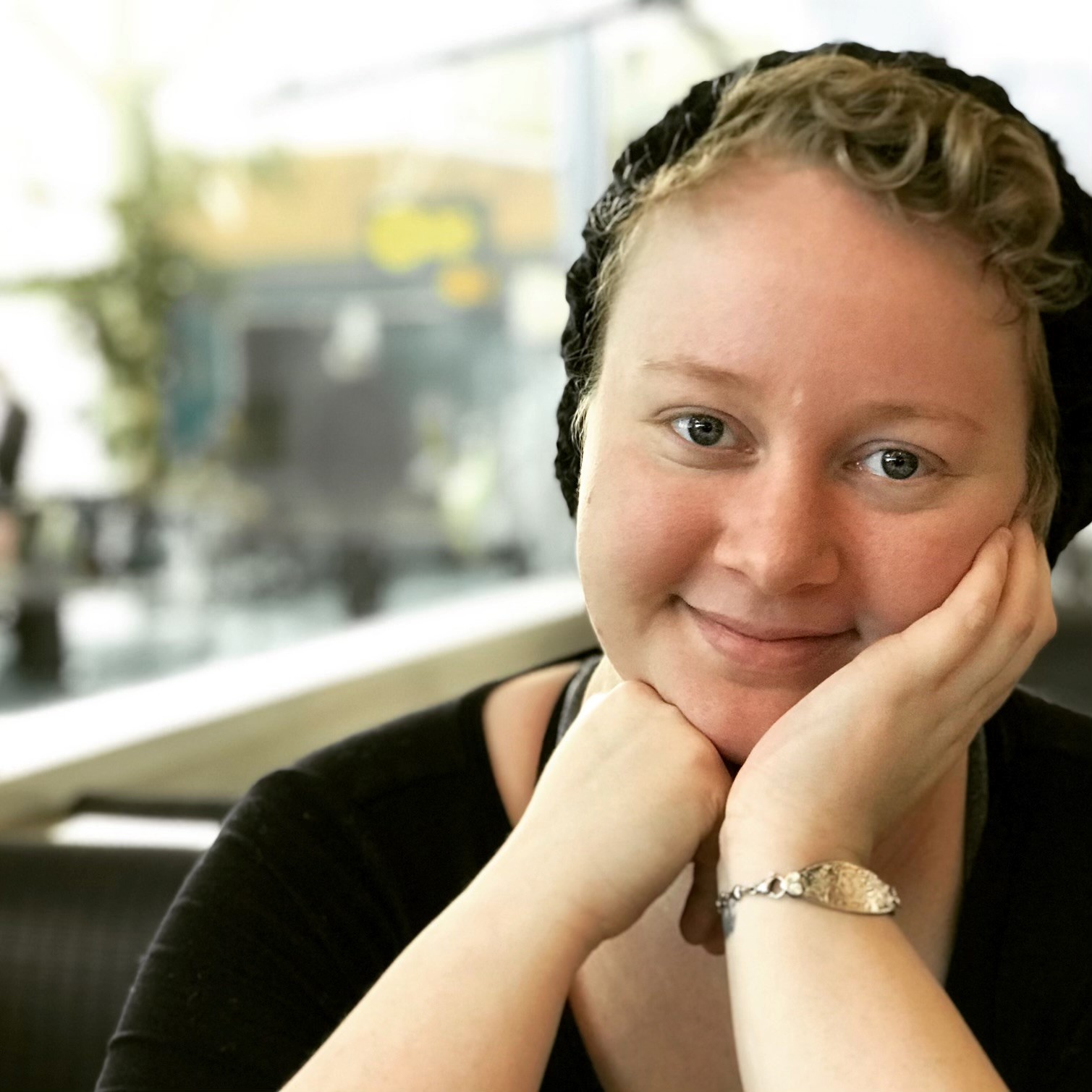 A person with short curly hair, wearing a black top, silver bracelet, and black head covering, smiles gently while resting their chin on their hand in a bright indoor setting at a Nanaimo Brain Injury Society event.