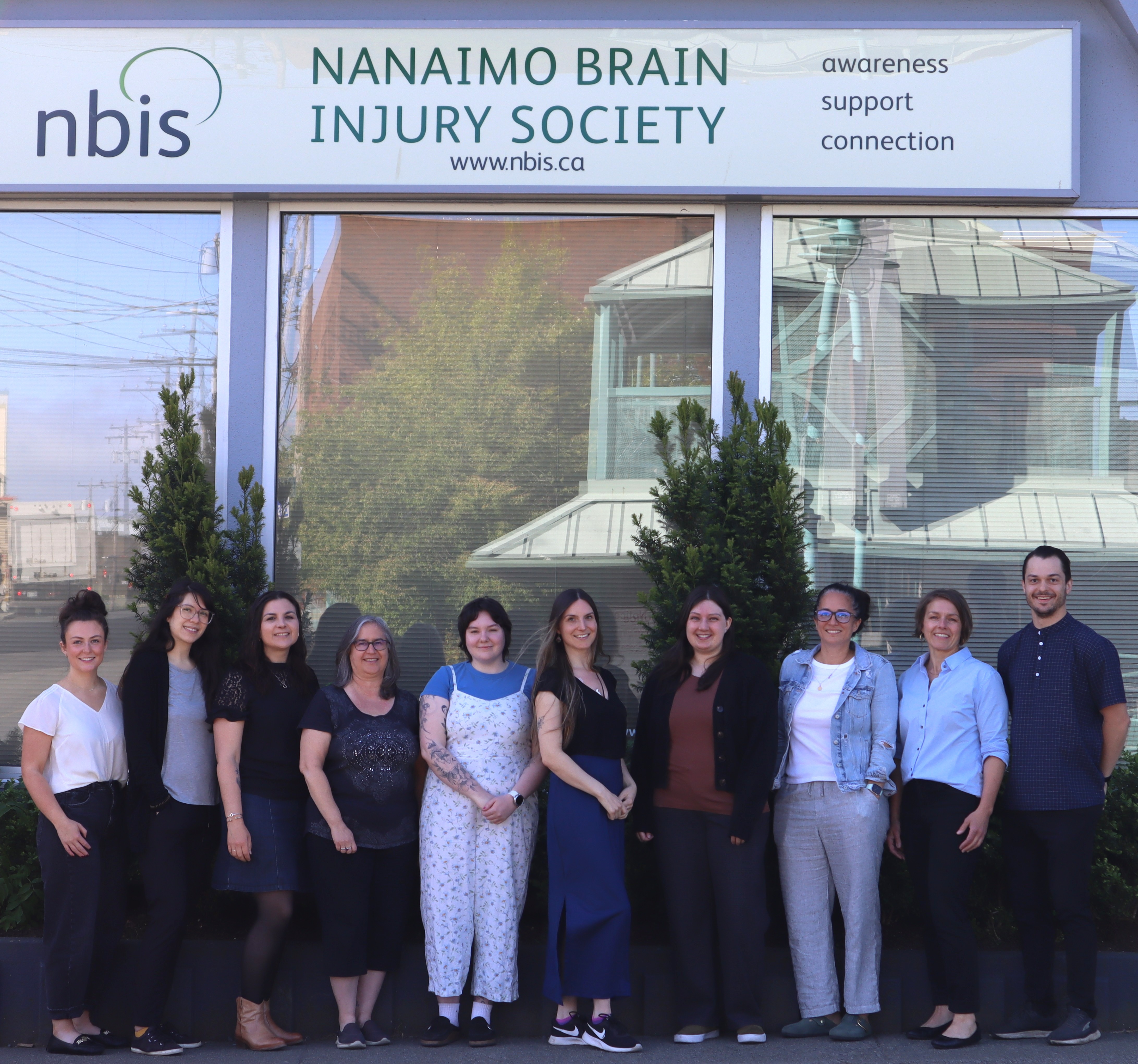 Ten people standing in a row and smiling in front of the Nanaimo Brain Injury Society building, with the organizations sign and logo visible above them.