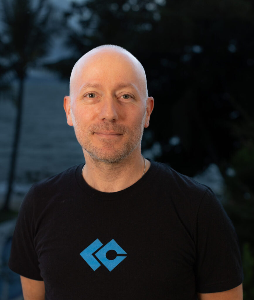 A man with a shaved head, wearing a black t-shirt with a blue geometric logo, stands outdoors with trees and water in the blurred background, representing the Nanaimo Brain Injury Society and Brain Injury Support.