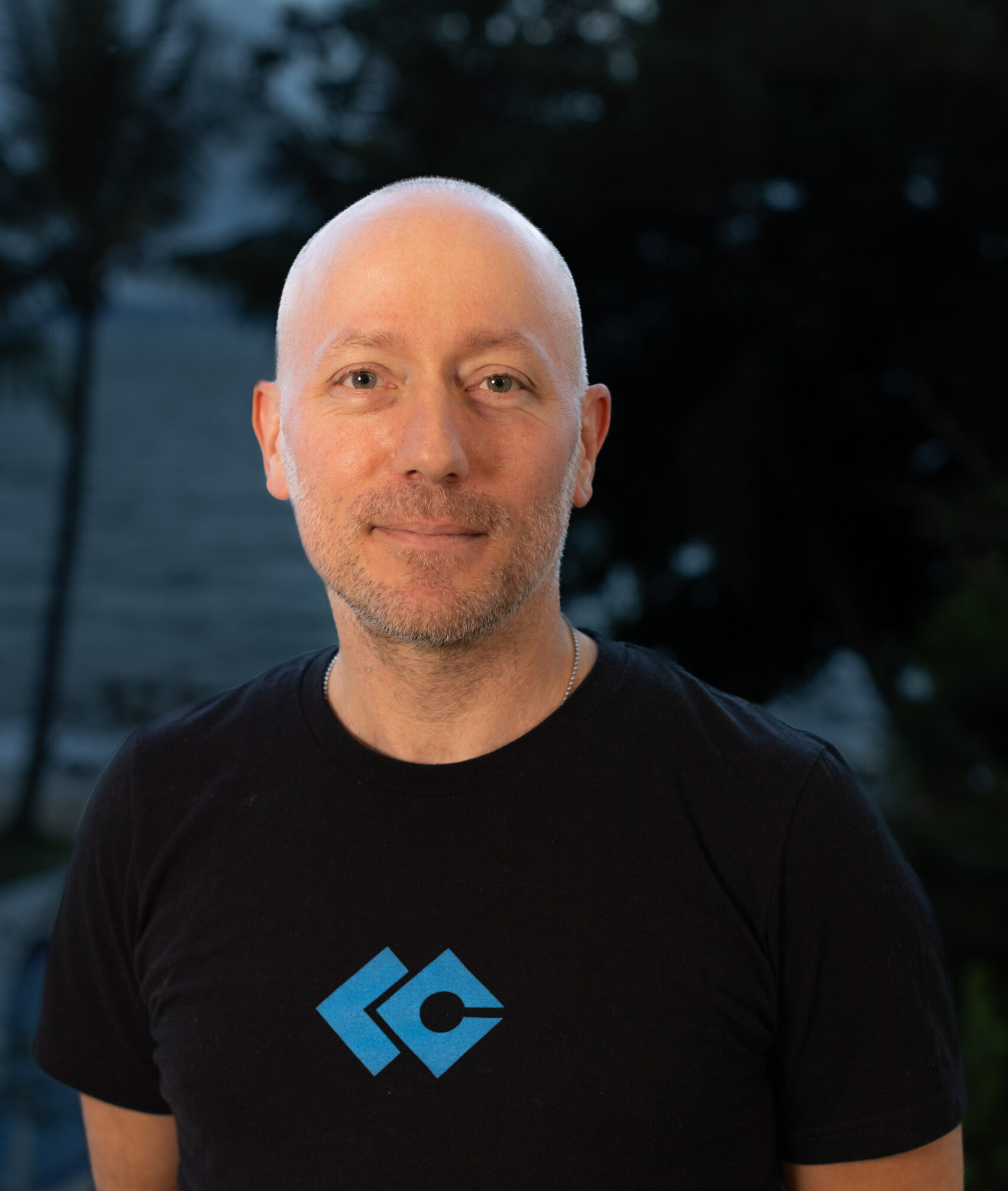 A man with a shaved head, wearing a black t-shirt with a blue geometric logo, stands outdoors with trees and water in the blurred background, representing the Nanaimo Brain Injury Society and Brain Injury Support.