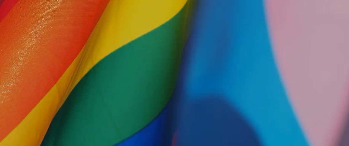 Close-up of overlapping rainbow and transgender pride flags, showing vibrant stripes of red, orange, yellow, green, blue, and pink—symbolizing inclusion and support from the Nanaimo Brain Injury Society (NBIS).