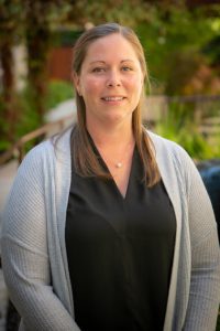 A woman with straight light brown hair, wearing a black blouse and light gray cardigan, smiles while standing outdoors with greenery in the blurred background, representing Nanaimo Brain Injury Society and their commitment to brain injury support.