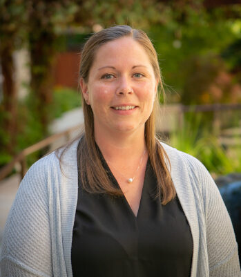 A woman with straight light brown hair, wearing a black blouse and light gray cardigan, smiles while standing outdoors with greenery in the blurred background, representing Nanaimo Brain Injury Society and their commitment to brain injury support.