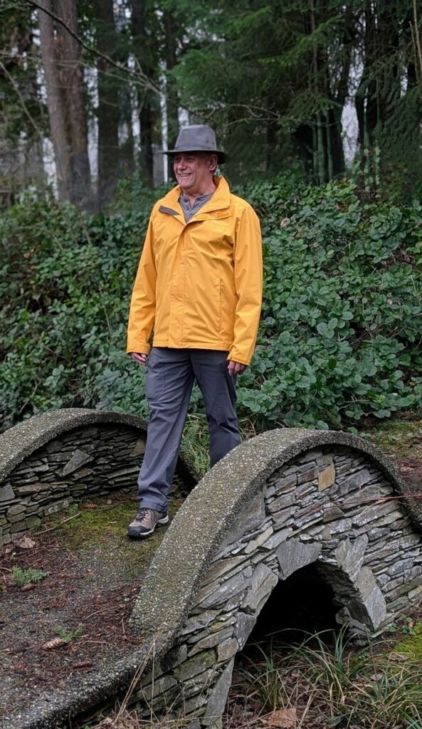 A man wearing a yellow jacket, gray pants, and a gray hat stands and smiles on a small stone bridge in a wooded area, embracing life after brain injury amid vibrant green foliage.