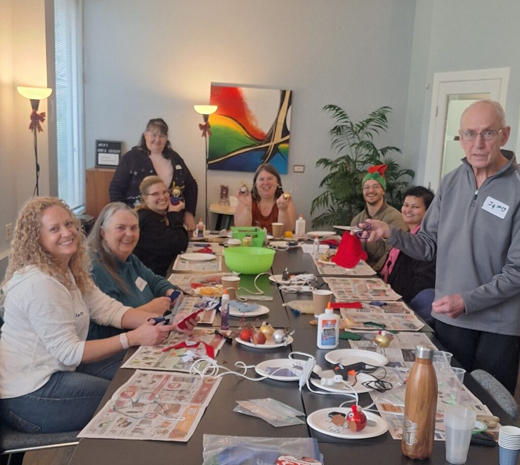 A group of people gather for a Christmas Crafting Session, sitting and standing around a table covered with craft supplies and newspapers, smiling at the camera. The brightly lit room is decorated with paintings, plants, and festive Jari accents.