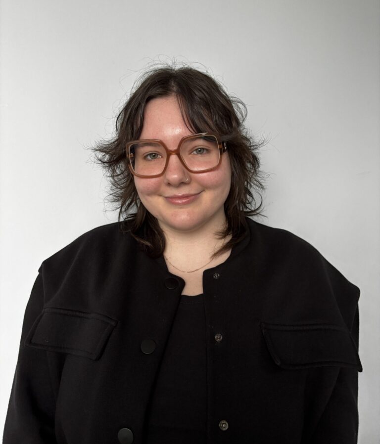 Anna Balaski, with shoulder-length brown hair and large brown glasses, smiles at the camera. She is wearing a black top and jacket, standing against a plain light gray background.