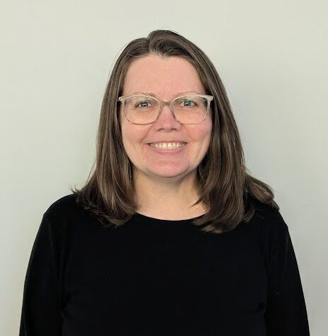Kate Jenkins, a woman with shoulder-length brown hair, wearing glasses and a black top, stands smiling in front of a plain white wall.