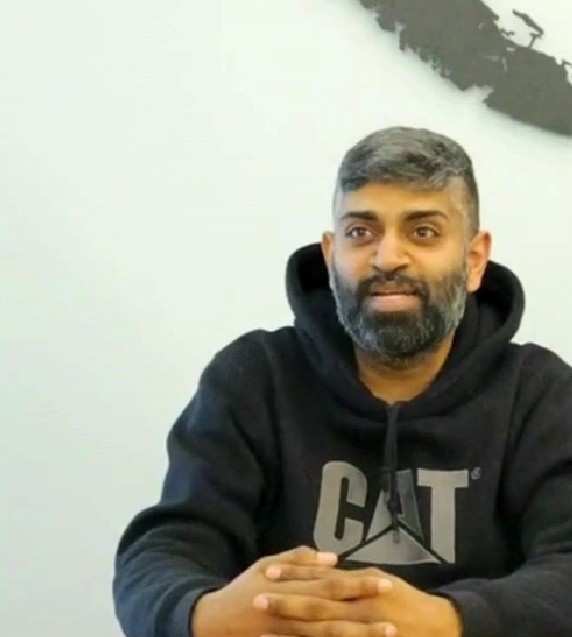 A man with short gray hair and a beard, wearing a black CAT hoodie, sits at a table with his hands folded. The calm setting hints at his role as a volunteer offering peer support. A light wall with a partial black design is in the background.