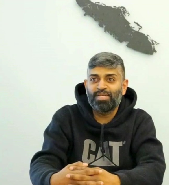 A man with short hair and a beard, wearing a black CAT hoodie, sits at a table with his hands folded, embodying the spirit of volunteer peer support. A black abstract wall art piece is visible on the light-colored wall behind him.