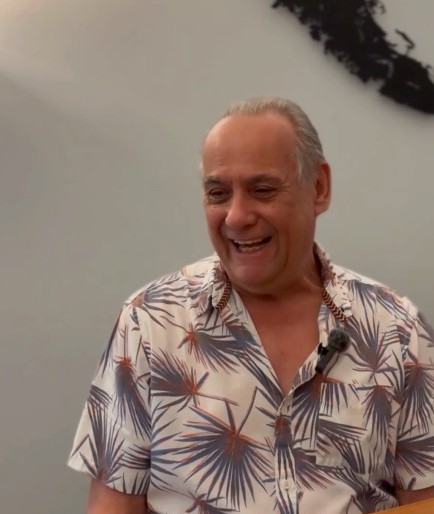 An older man with gray hair is smiling and wearing a patterned short-sleeve shirt. A volunteer in Peer Support, he has a microphone clipped to his collar and is sitting indoors against a plain, light-colored wall.