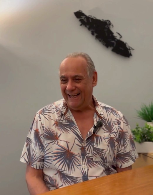 A smiling older man in a patterned, short-sleeved shirt sits at a wooden table, embodying the spirit of volunteer peer support. Behind him, a small plant and a black abstract wall decoration are visible on a light-colored wall.